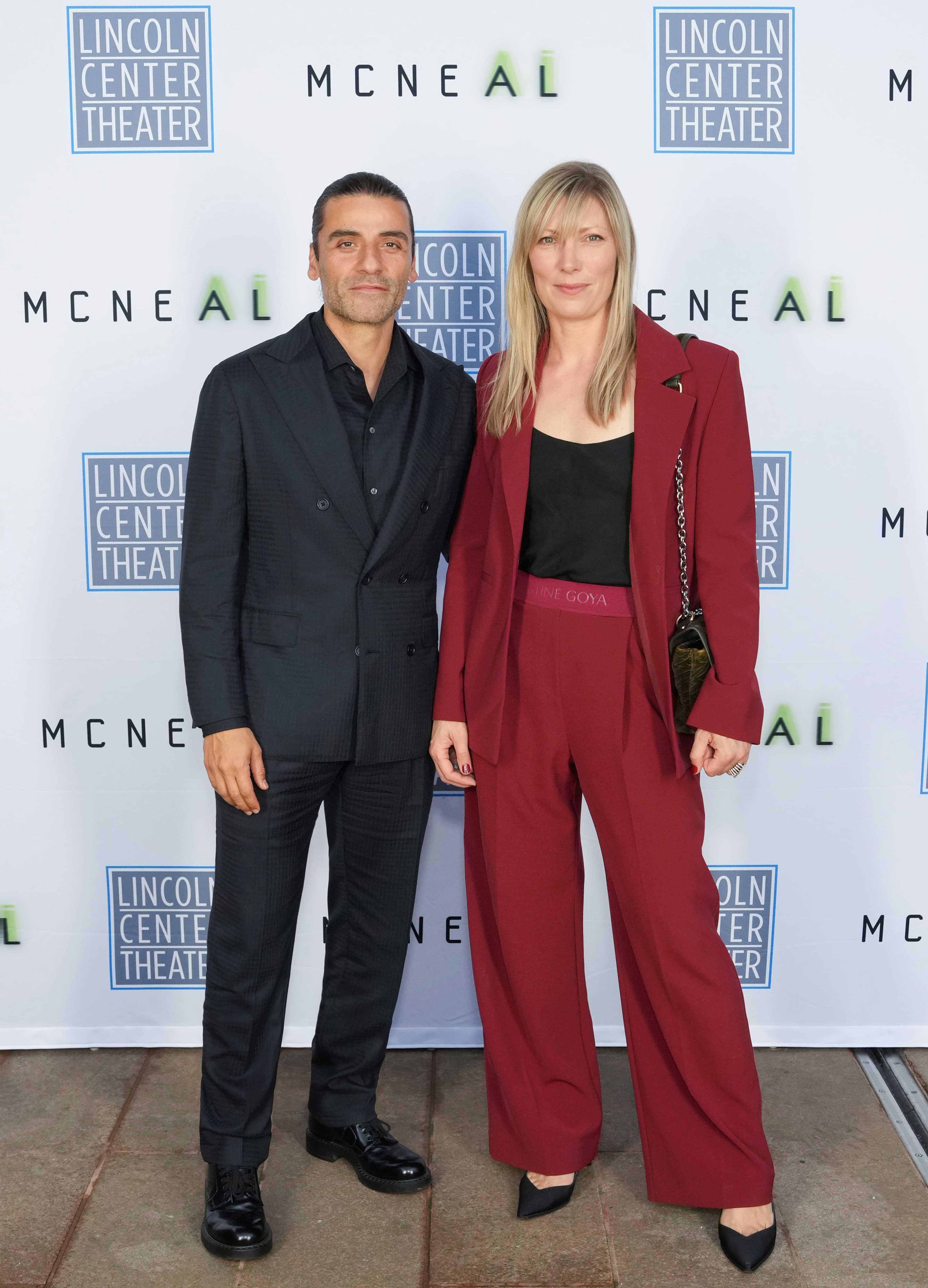 Oscar Isaac at Lincoln Center Theater event - man in dark suit and woman in burgundy pantsuit against branded backdrop.