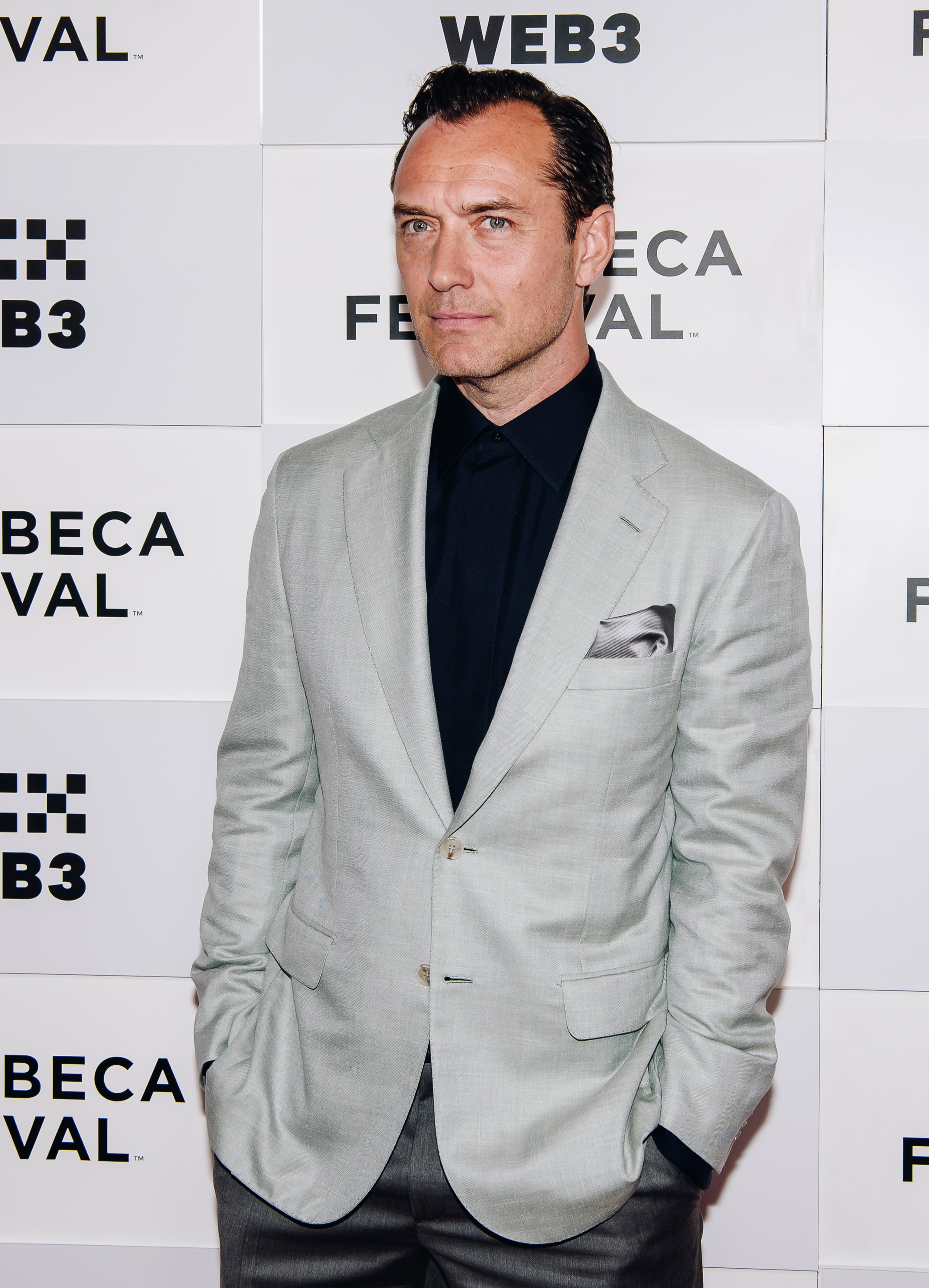 Jude Law in Brioni light gray blazer and navy shirt at Tribeca Festival event, standing against festival logo backdrop.