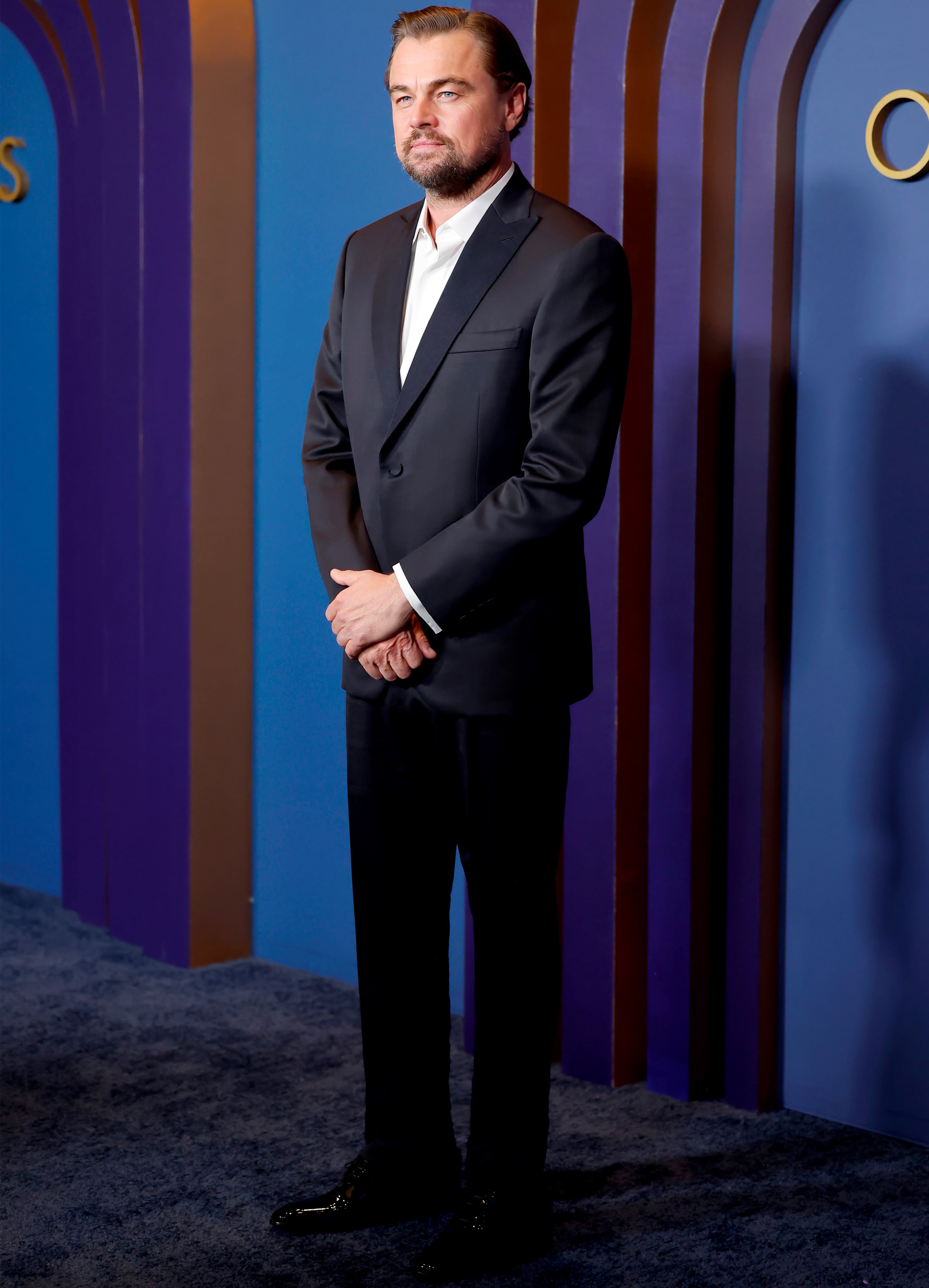 Leonardo di Caprio in Brioni black suit and white shirt standing at a formal event with blue and purple striped backdrop.
