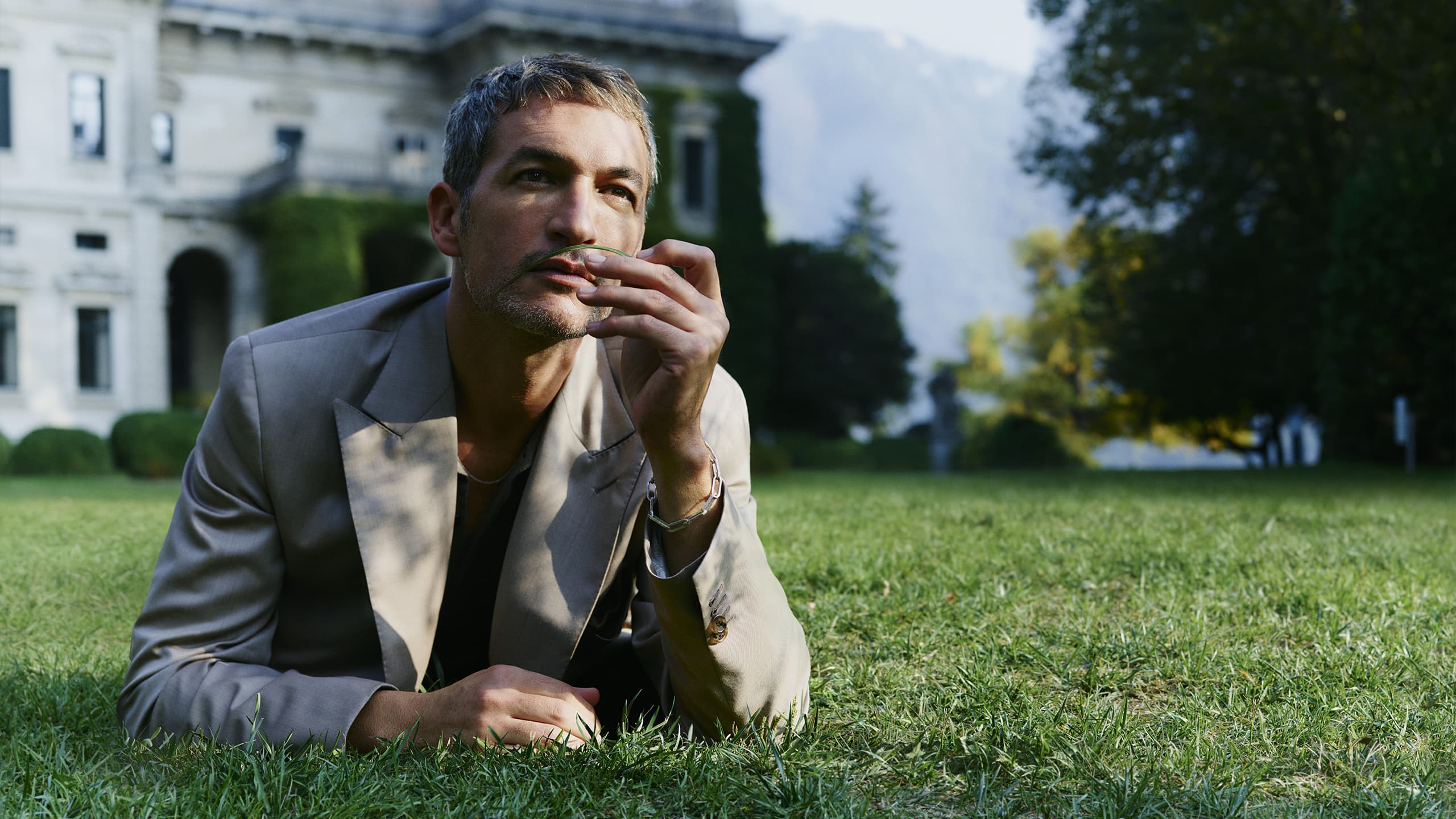 Male model in light suit lying on grass, thoughtfully gazing upward with hand near face; elegant mansion in background.