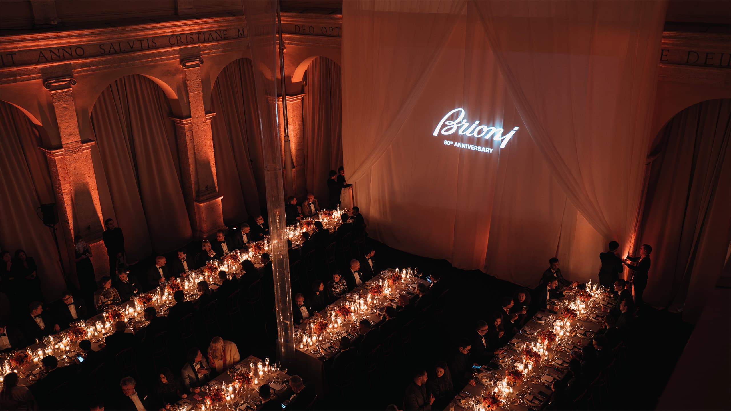 Elegant Brioni 80th anniversary gala dinner in historic venue with candlelit tables and glowing logo on draped wall.