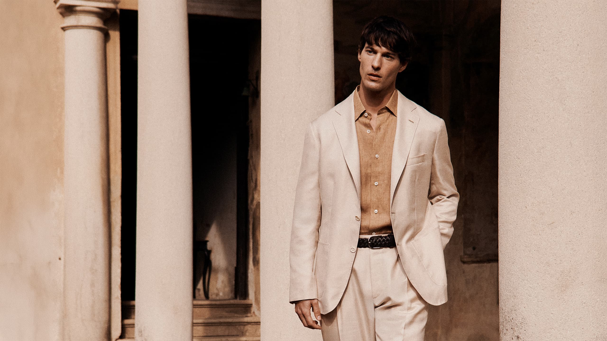 Young man in Brioni light blue blazer and beige pants standing against weathered beige and brown textured wall with natural lighting.