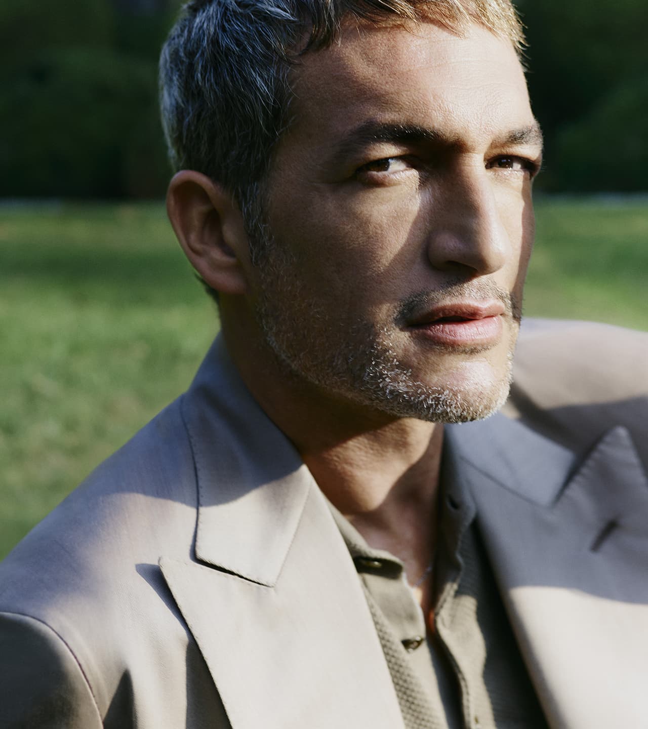 Close-up portrait of a male model with short gray-streaked hair wearing a light beige suit against a green outdoor background.