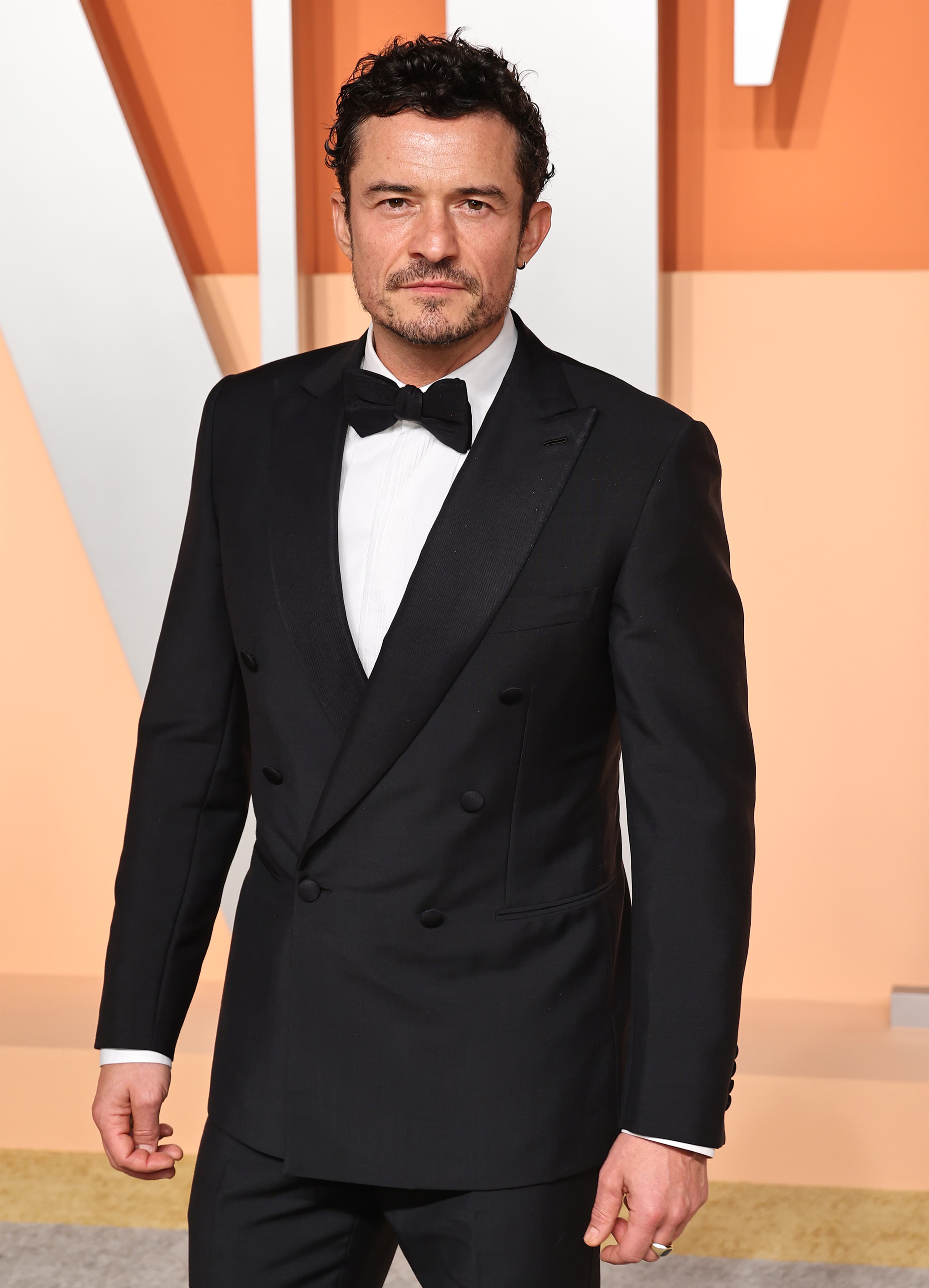 Orlando Bloom in elegant Brioni black tuxedo with bow tie at formal event against peach-colored backdrop.