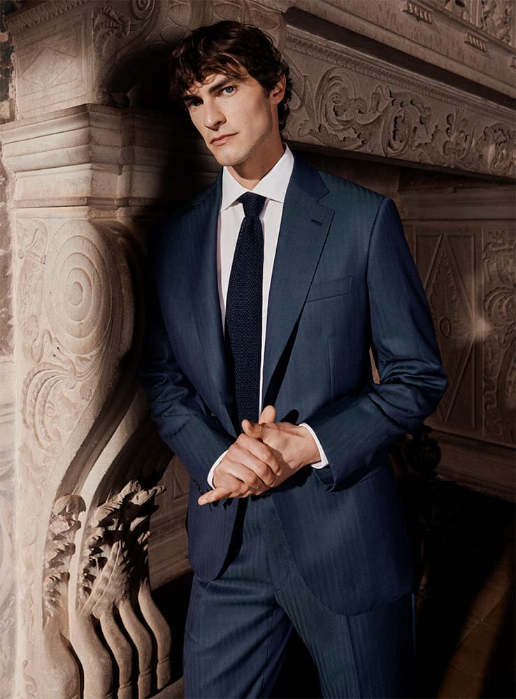 Young man in Brioni navy blue suit with white shirt and dark tie standing against ornate stone architectural columns and carvings.