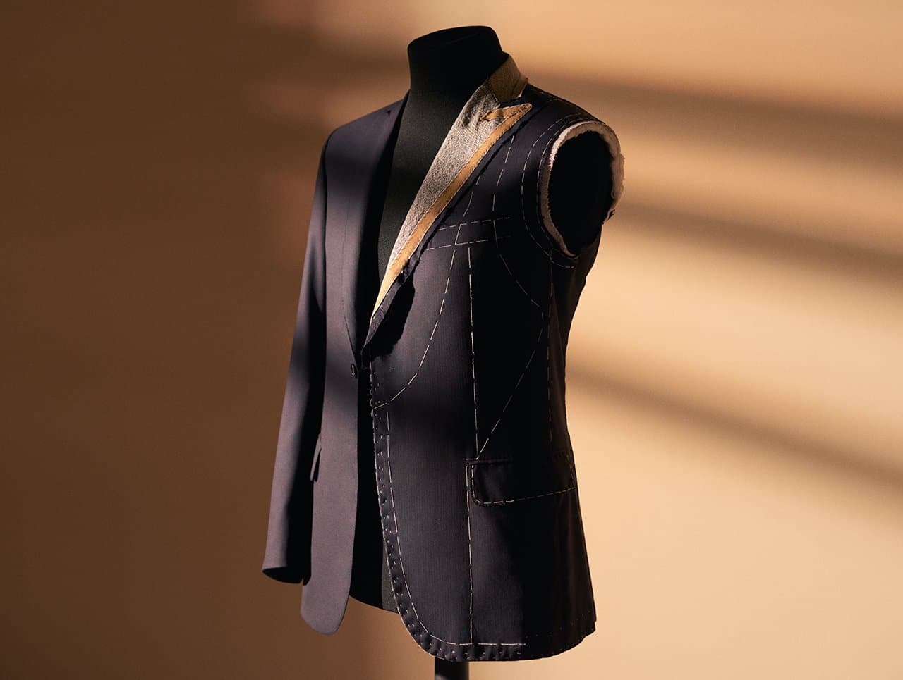 Navy blue tailored Brioni suit jacket on a mannequin showing basting stitches and construction details against warm backdrop.