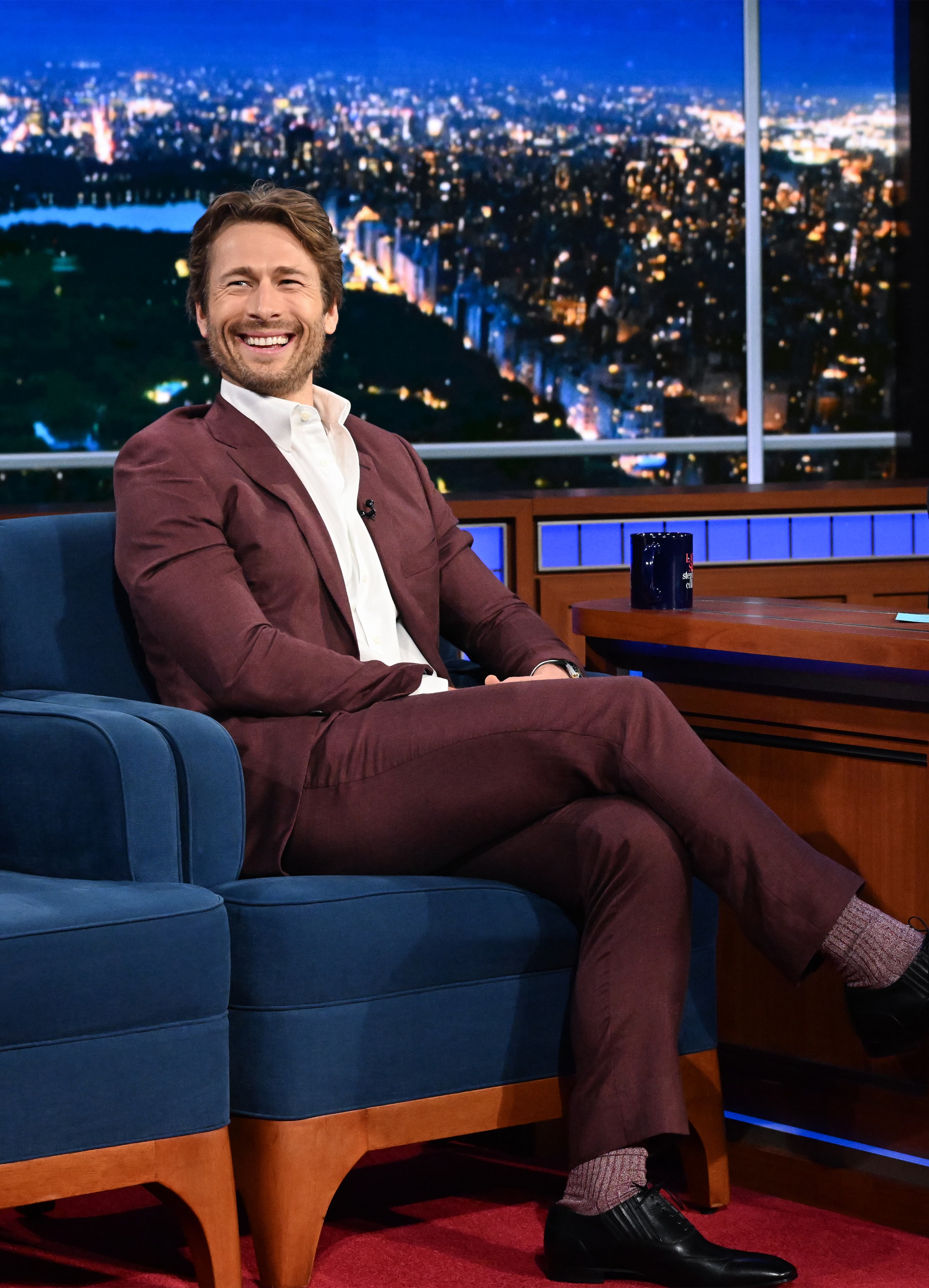 Glen Powell in burgundy Brioni suit smiling on talk show set with nighttime city skyline visible through window behind.