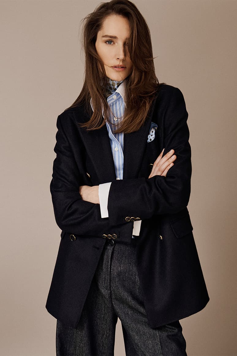 Woman in Brioni black blazer with blue striped shirt and dark trousers, arms crossed, against beige background in professional portrait.