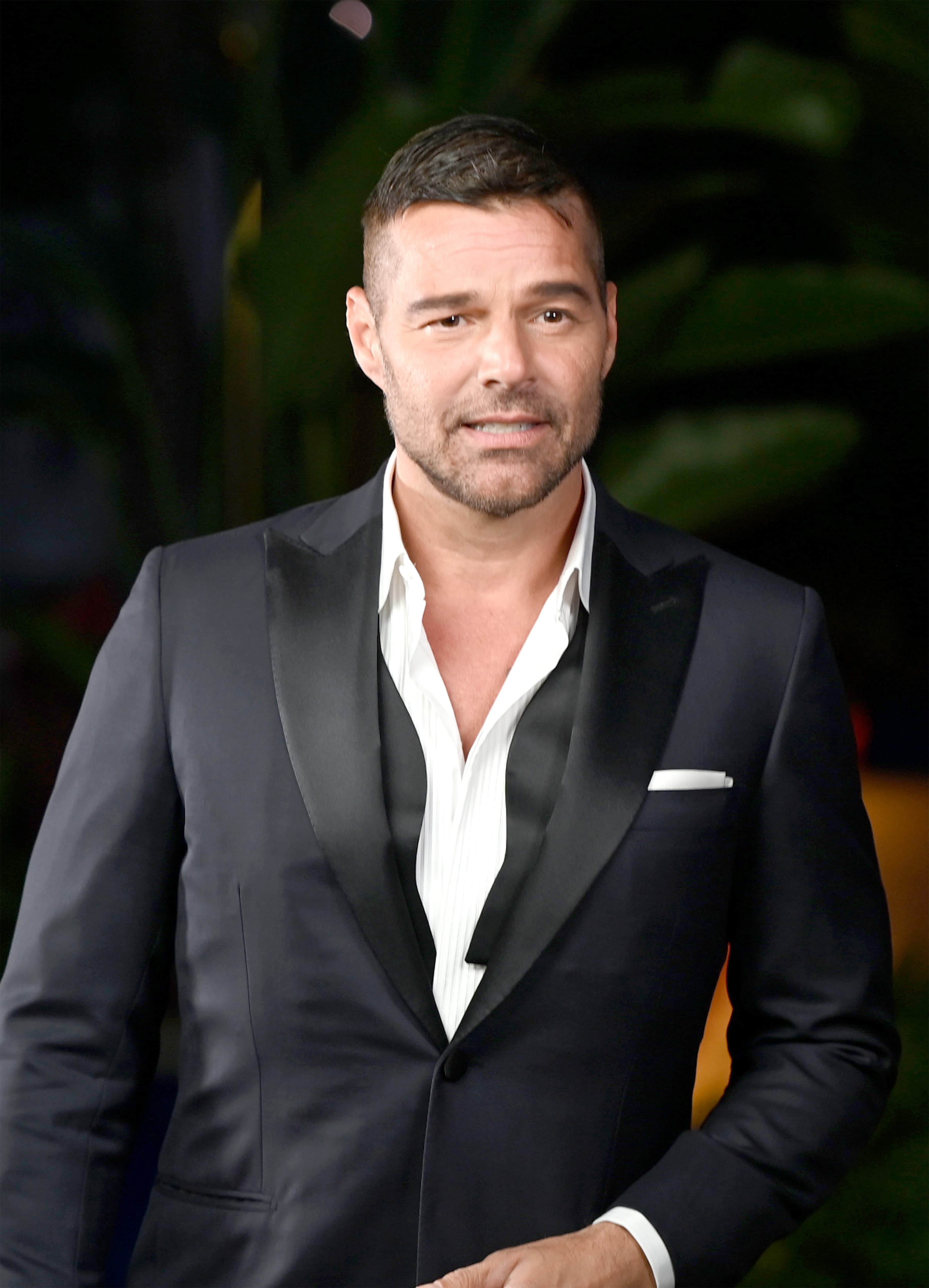 Ricky Martin in elegant Brioni black tuxedo with white shirt at formal event against dark green background.