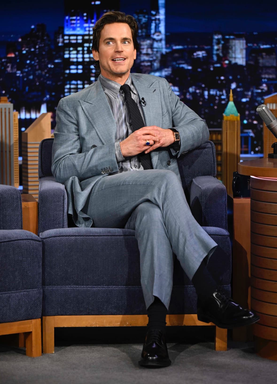Matt Bomer in Brioni gray suit sitting on blue armchair during late-night talk show interview with city skyline backdrop.