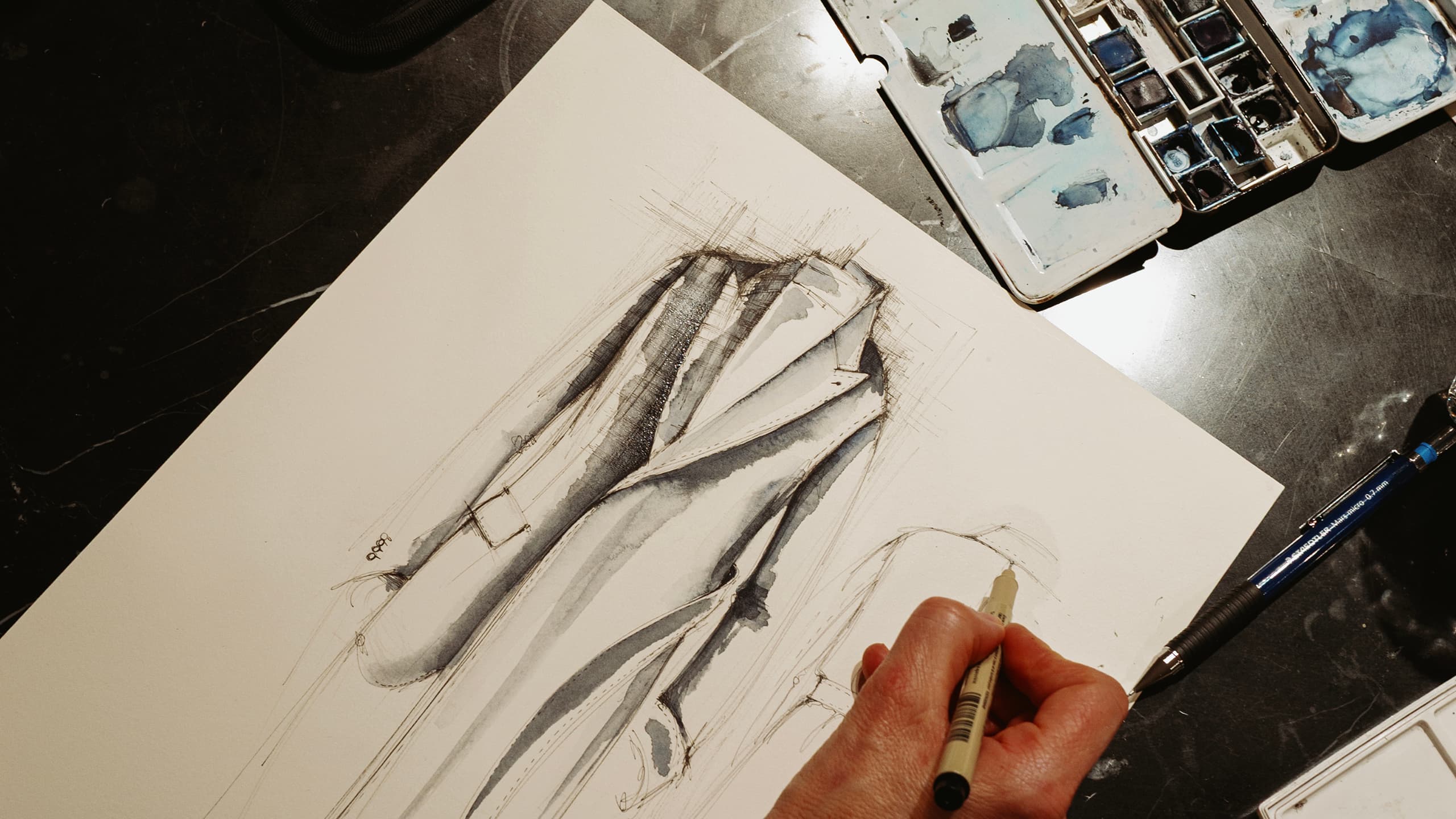Artist's hand holding brush over watercolor sketch of draped fabric in brown and gray tones, with paint palette nearby.