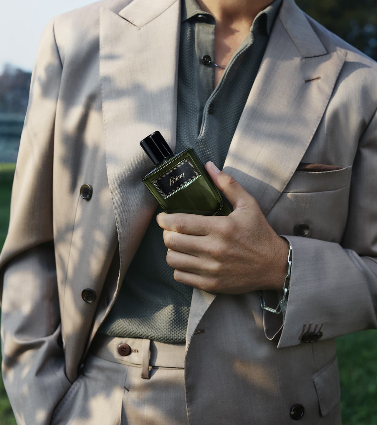 Male model in beige suit holding Essentiel green bottle against their chest in outdoor lighting.
